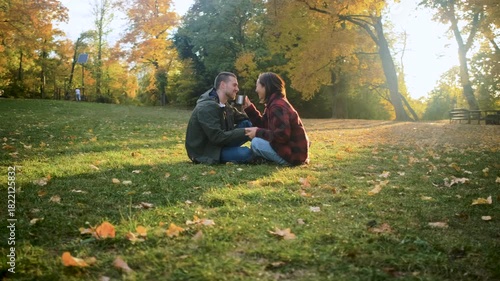 Romantic young multiracial couple enjoying a date in an autumn park, sitting on grass drinking tea.