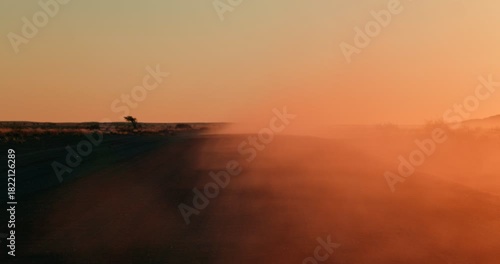Orange dust rises over a gravel road cutting through Namibia’s red desert at sunset, with warm glowing light reflecting beautifully in the drifting airborne dust.