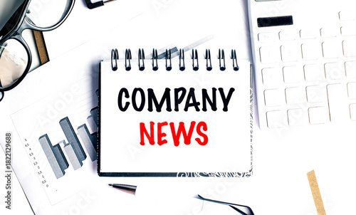 Company news phrase written on notepad with glasses, pen, charts and keyboard on white desk background
