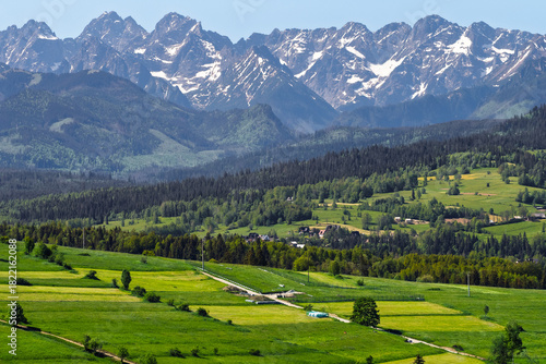 Fototapeta Naklejka Na Ścianę i Meble -  Panoramic view of the snow-capped Tatra Mountains from Rzepiska village in Podhale, Poland. Scenic June landscape with vivid green fields and rural settlement.