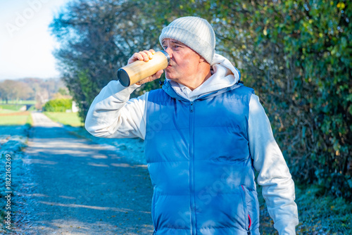 senior male runner in sportswear taking break during morning run drinking warm beverage, hot drink from wooden thermos, self care and wellness, feeling energized, outdoors healthy and active lifestyle