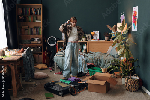 Konstfotografi Caucasian teenage girl standing in messy bedroom packing suitcase and cardboard