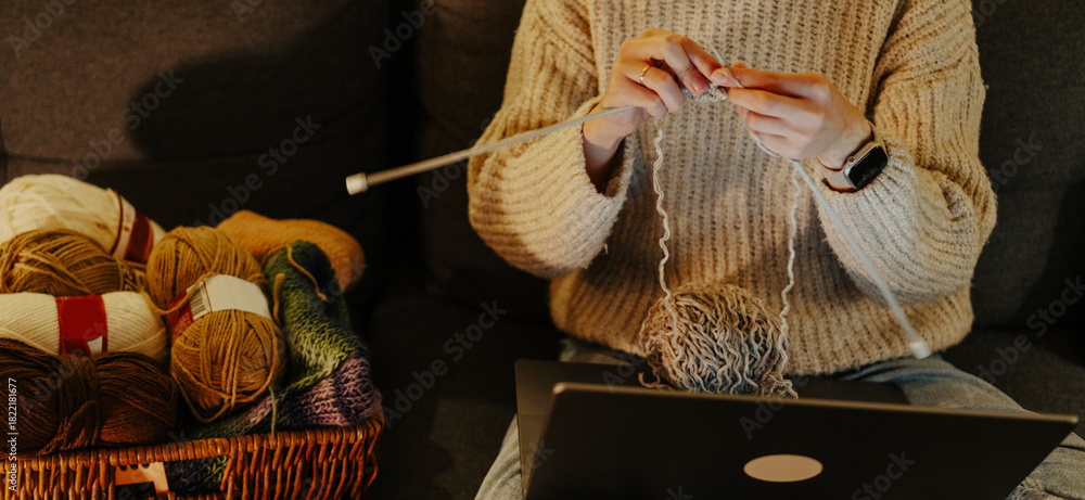 Fototapeta premium A woman knits while sitting on a sofa, with a basket of yarn nearby.