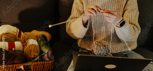 A woman knits while sitting on a sofa, with a basket of yarn nearby.