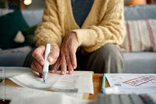 Senior woman reviewing invoices and bills at home, holding pen and analyzing financial documents on table, close up of hands and paperwork, managing personal finances