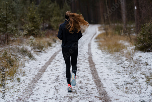 Focused on the path ahead: A rear view of a young woman running through a cold, snowy forest road, symbolizing moving forward and achieving goals.