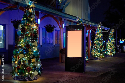 Fototapeta Naklejka Na Ścianę i Meble -  Multiple small Christmas trees with shiny ornaments line the path near a storefront. A blank digital sign stands among the glowing holiday lights.