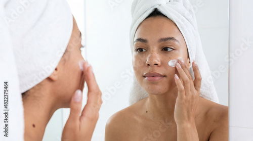 A young woman with natural skin and freckles applies moisturizing cream to her cheek while looking at her reflection in a bathroom mirror, wearing a white towel on her head.