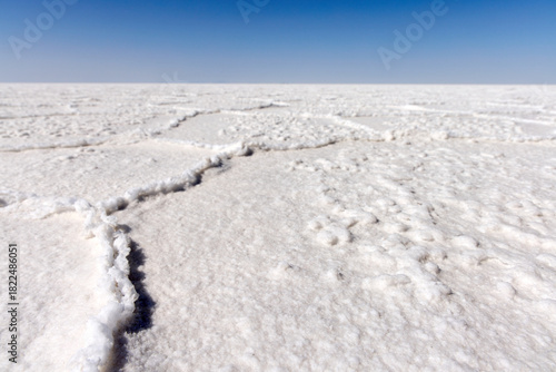 A view across the cracked, seemingly endless white salt crust of Salar de Uyuni. A clear blue sky dominates above the horizon line of the salt flat expanse.