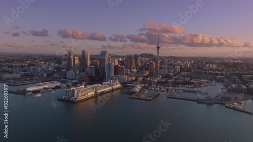 Aerial view of Auckland, New Zealand, at sunset. The city skyline features modern buildings and the iconic Sky Tower. The harbor is filled with boats.