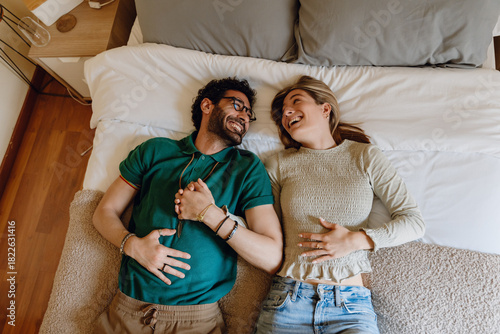A man and a woman are holding hands and lying on a bed while laughing and looking at each other