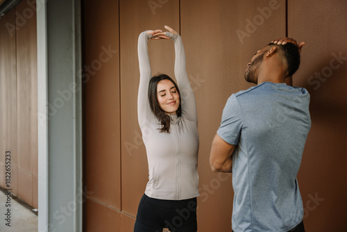 A female athlete stretches her arms above her head with her eyes closed while standing opposite a male athlete who stretches his neck and tilts his head to the side