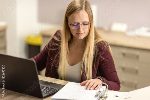 Woman working from home with laptop and paperwork, focused on organizing documents at her desk