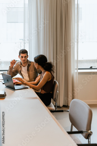 A male worker holds a pencil and listens to a female worker sitting next to him at a desk and holding her hands over a laptop
