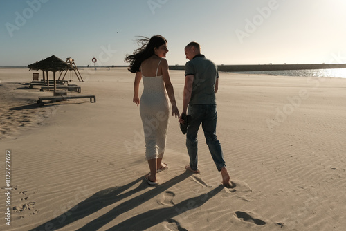 Couple walking barefoot on sandy beach near ocean at sunset, wind blowing her hair in a peaceful romantic scene.
