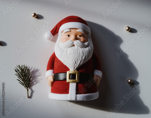 Santa claus figurine sitting on white surface surrounded by gold beads and pine sprig creating festive holiday scene with bright lighting.