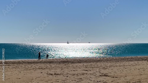 Two silhouetted figures walk along the shoreline, facing a dazzling expanse of sunlit water that sparkles brightly across the deep blue sea. Two small sailboats are visible on the distant horizon.