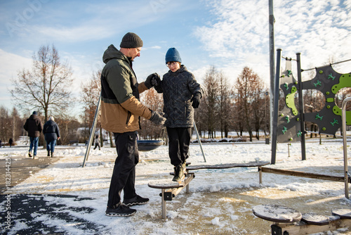 Father helping a child balance on playground stepping stones in a snowy park on a clear winter day, capturing a moment of outdoor play, trust and learning.
