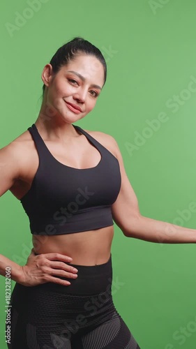 Vertical clip. Young sportswoman shows off her physical strength by lifting dumbbells on green background in studio.