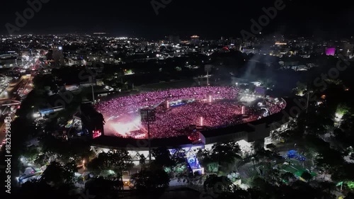 Aerial approaching shot showing crowd of people celebrating bad bunny festival show in open air stadium at night. Santo Domingo, Dominican Republic. Music show at night with flashing illuminated light