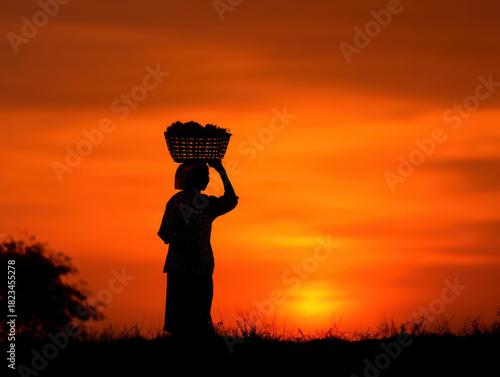 Wallpaper Mural Silhouette of person carrying basket on their head is set against vibrant orange sunset sky, creating peaceful and serene scene. image captures essence of rural life and beauty of nature Torontodigital.ca