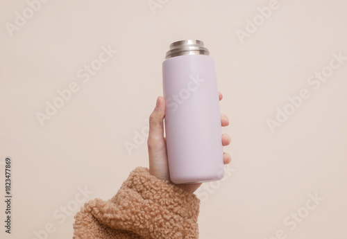 Woman's hand in a cozy sherpa sweatshirt holds metal thermos bottle on a beige background