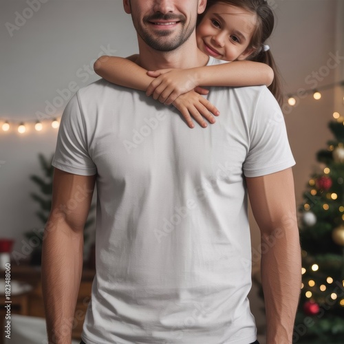 Handsome male model in slim‑fit white tee standing in Christmas‑decorated room, hugged from behind by little girl around his neck. Men’s basic t-shirt mockup, father and daughter, family indoors