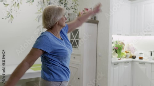 Senior woman performing targeted arm circles and stretching exercises, maintaining physical mobility and fitness within home kitchen environment, demonstrating active aging and wellness commitment