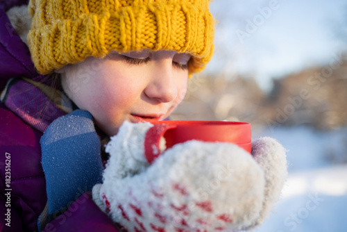 A child drinks hot tea from a thermos cup outdoors in a park during the Christmas holidays in winter.