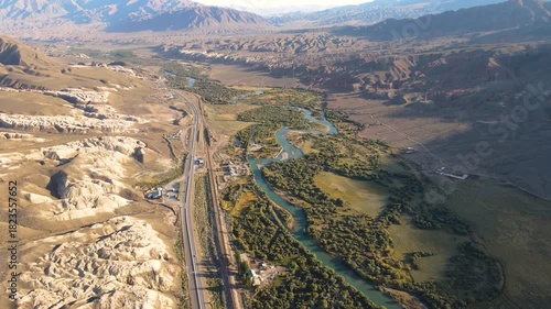 Sweeping aerial view of a mountain valley showing a highway and railroad running parallel to a turquoise river, flanked by green riparian areas and sculpted badlands