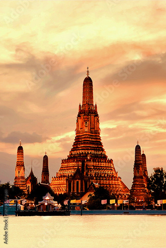 Wat Arun glows in golden light at sunset, reflecting over the Chao Phraya River under a dramatic evening sky in Bangkok, Thailand.