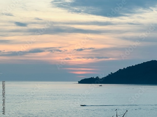 A peaceful ocean scene at sunset with a small boat passing in front of distant mountains under an orange sky. Calm, dreamy and warm evening nature landscape.