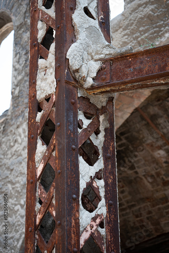 Close-up of a rusted metallic lattice column and beam junction in an abandoned factory, showing exposed rivets, heavy corrosion, and broken cement coating.