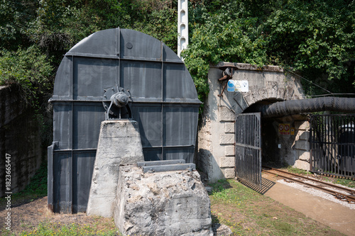 Exterior of a giant, historical black centrifugal fan by Victor Gruber next to the gated, abandoned coal mine tunnel entrance, surrounded by greenery.