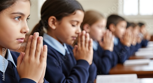 Group of children with clasped hands in prayer, wearing navy blue uniforms, engaged in a moment of reflection, embodying the spirit of faith and community in a serene classroom environment