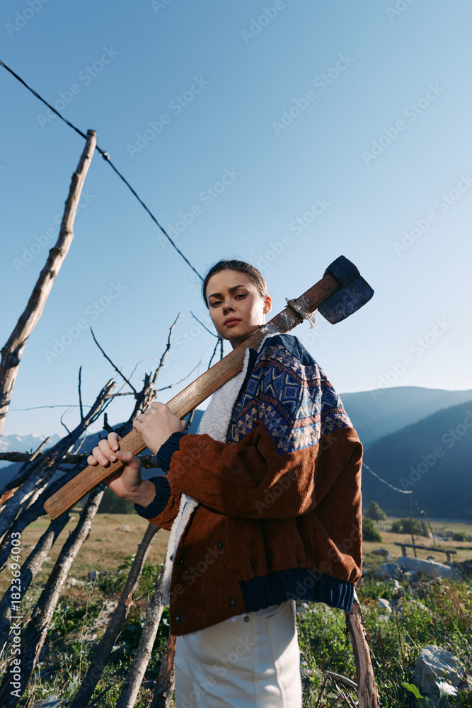 Fototapeta premium Woman with axe over shoulder by wooden fence in rural mountains landscape, outdoor portrait of a strong farmer or gardener in warm sweater, countryside scene with clear sky.