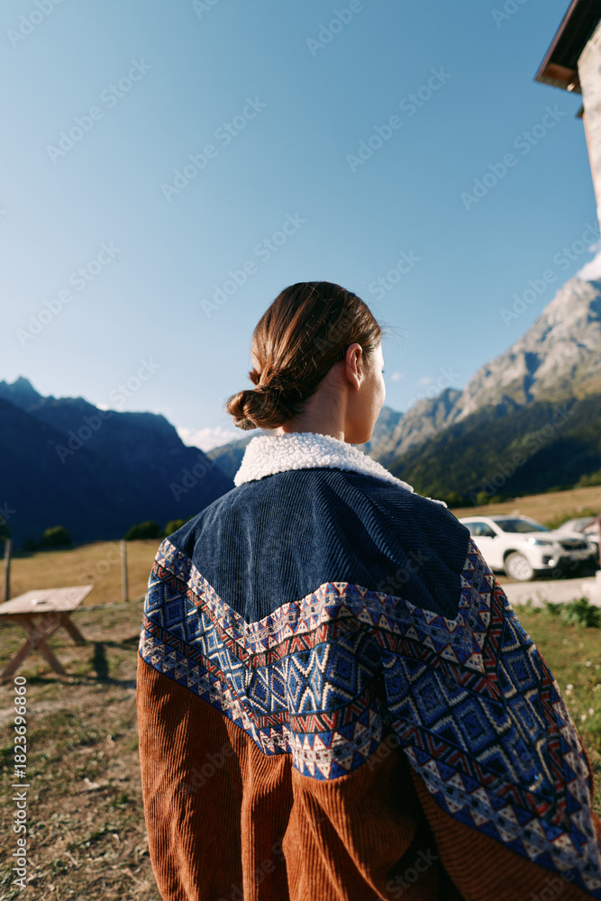 Fototapeta premium Woman in a patterned sweater stands with her back to camera in the mountains outdoors, overlooking a quiet countryside landscape under clear sky, enjoying autumn light and peaceful nature.