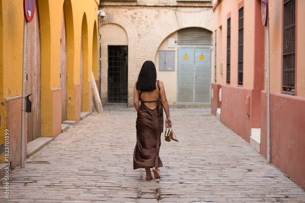 Φωτογραφία A young, dark-haired woman wearing a brown dress and holding gold heels in her hand, with her back turned, walks barefoot down a lonely alleyway