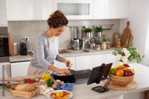 Fotografie A woman prepares a nutritious dish in a sleek kitchen, surrounded by fresh vegetables and fruits, while following a recipe on a tablet device