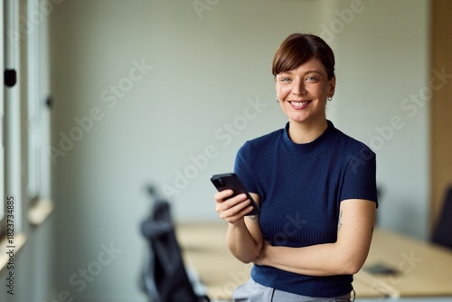 Confident Woman With Smartphone in Modern Office Workspace, Smiling While Standing by Desk