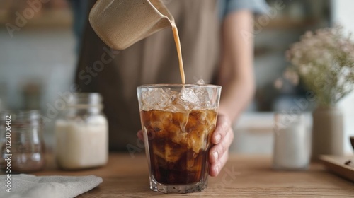 Person's hand holding a glass of iced coffee. the glass is filled with ice cubes and the coffee is being poured from a beige-colored pitcher into it.