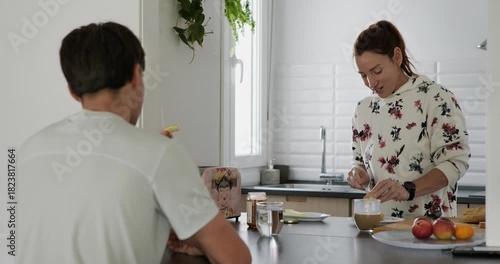 A Caucasian woman is spreading butter or jam on toast while talking to a young Caucasian man who is sitting opposite her at the kitchen counter.