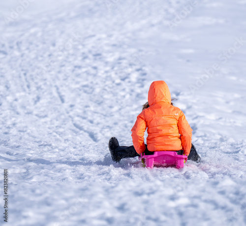 A child in a vibrant orange winter jacket sleds down a pristine white snowy hill on a bright, sunny day. Focus on the joy of winter outdoor activities.