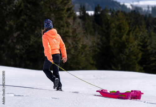 A child in a bright orange jacket and winter hat walks through deep, fresh snow, pulling a pink sled. Forested mountains create a serene winter wonderland background.