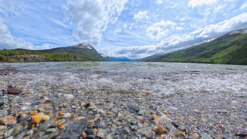 Peaceful lake in Parque Nacional Tierra del Fuego, Argentina, with small waves lapping the rocky shore. In the distance, majestic mountains rise under a calm sky, creating a tranquil landscape.
