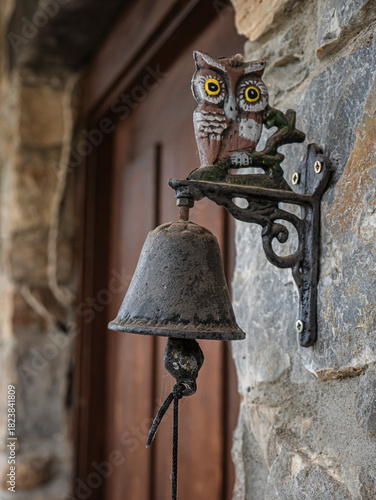 A ceramic owl rests on the stand of a small bronze bell (old hearth doorbell) with a wooden door behind it in a stone house in a rural setting