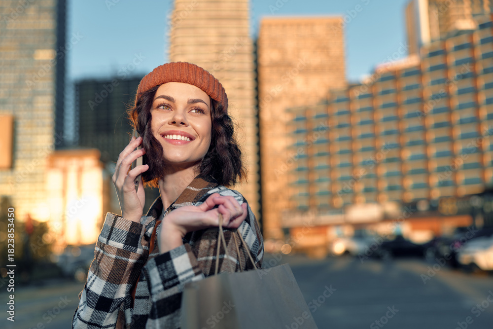 Fototapeta premium Woman shopping on city street, phone to ear and smile, beanie and bag in hand in warm sunlight; authenticity and candid lifestyle portrait with golden glow and emotional storytelling.