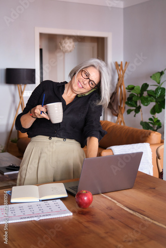 Smiling mature businesswoman working from home, drinking coffee and talking on the phone