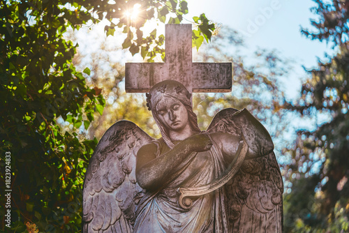 Angelic Statue With Cross and Wings in Sunlit Cemetery Garden