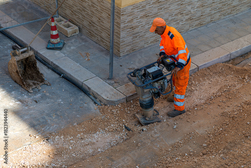 Construction worker in orange safety gear operating a tamping rammer machine to compact gravel in a trench. Road maintenance scene with an excavator bucket and traffic cone nearby.